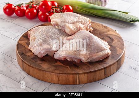 Rohe Hähnchenschenkel auf rundem Holzschneidebrett angeordnet. Hellrote Kirschtomaten grüner Lauch im Hintergrund. Saubere weiße Fliesenoberfläche. Natürliche Farbe Stockfoto