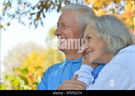 Senior umarmt und lächelt in einer herbstlichen Außenszene mit gefallenen Blättern und Bäumen, Nahaufnahme seitlich, blaue und graue Pullover, glücklich und Stockfoto