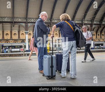 Zwei Passagiere mit Koffern unterhalten sich auf einem Bahnsteig. Andere Passagiere gehen vorbei und ein Zug mit einer Reihe von Waggons steht im Hintergrund Stockfoto