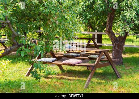Gemütlicher Picknick- und Entspannungsbereich mit Holztisch unter schattigen Bäumen. Outdoor-Café-Atmosphäre in einem grünen Garten, perfekt für Freizeit, Reisen, Familienga Stockfoto