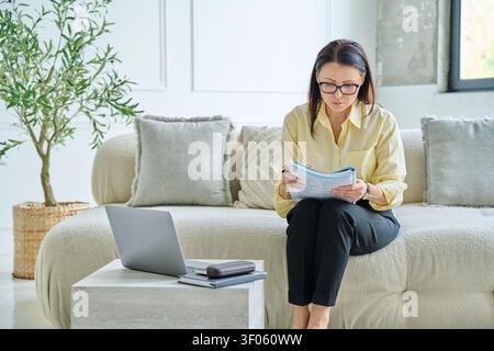 Konzentrierte reife Geschäftsfrau liest Dokument mit Laptop auf dem Sofa zu Hause. Stockfoto