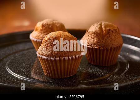 drei frisch gebackene Muffins auf schwarzem strukturiertem Teller, goldbraune Rührkuchen in Papierbechern, Seitenansicht mit weichem Fokus und Kopierraum, warm hausgemacht Stockfoto