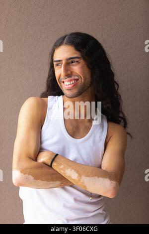 Ein lächelnder Mann aus dem Nahen Osten mit Vitiligo steht mit überkreuzten Armen, trägt ein weißes Tanktop an einer neutralen Wand. Hochlichtmarker mit natürlichem Licht Stockfoto