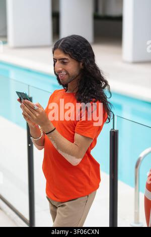Ein Mann aus dem Nahen Osten mit Vitiligo steht an einem modernen Swimmingpool, lächelt, während er ein Smartphone benutzt, trägt ein orangenes Hemd bei hellem Tageslicht und entspannt sich Stockfoto