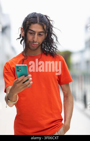 Porträt eines Mannes aus dem Nahen Osten mit Vitiligo, der sein Smartphone überprüft, während er draußen auf einer Stadtstraße läuft, ein oranges T-Shirt trägt und schaut Stockfoto