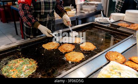 Ein Straßenverkäufer auf dem Gukje Market, Busan, bereitet gekonnt bindaetteok, traditionelle koreanische Mungbohnen-Pfannkuchen, auf einer heißen Grillplatte zu. Eine lebendige Szene aus Stockfoto