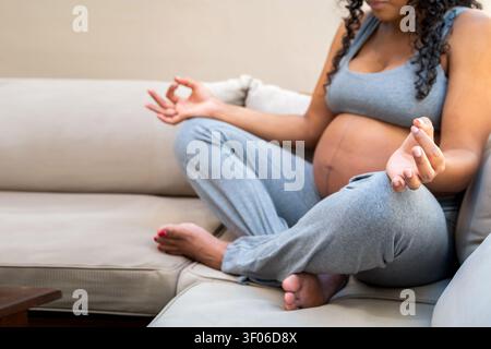 Nahaufnahme einer schwangeren Frau, die im Kreuz auf einem Sofa sitzt, Meditation mit entspannten Händen und ruhigem Atmen praktiziert, Wohlbefinden fördert, achtsam Stockfoto