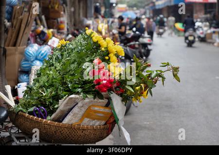 Eine belebte Hanoi Straße zeigt eine lebhafte Ausstellung von bunten Blumen in einem gewebten Korb, die das lebendige Wesen und den alltäglichen Charme der Stadt einfangen Stockfoto