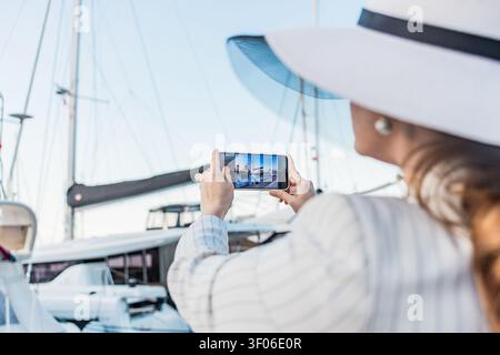 Elegante weibliche Touristin mit breiter Krempe fotografiert Luxusyachten in einem sonnigen Yachthafen mit einem Smartphone, um Reiseerinnerungen und KüstenV festzuhalten Stockfoto