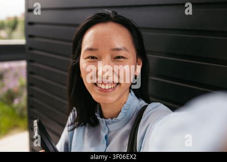 Lächelnde asiatische Geschäftsfrau, die im Freien ein Selfie mit ihrem Smartphone macht, hält einen selbstbewussten und freudigen Moment fest. Sie trägt professionelle Anzüge Stockfoto