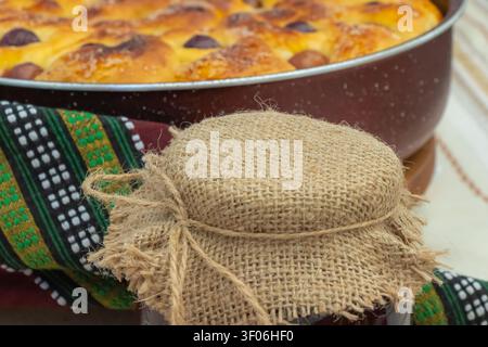 Hausgemachte Fruchtmarmelade in einem rustikalen mit Sackleinen bedeckten Glasgefäß mit frisch gebackenem süßem Brot im Hintergrund, das ein traditionelles hausgemachtes Frühstück erschafft Stockfoto