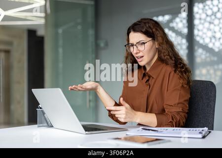 Junge Geschäftsfrau mit Brille hat ein Problem und sieht frustriert aus, während sie in einem modernen Büro an einem Laptop-Computer arbeitet Stockfoto
