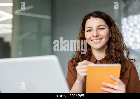 Junge berufstätige Frau lächelt und schaut auf einen Laptop-Bildschirm. Hält ein orangefarbenes Notizbuch und einen Stift. Macht Notizen in einer hellen modernen Büroumgebung Stockfoto