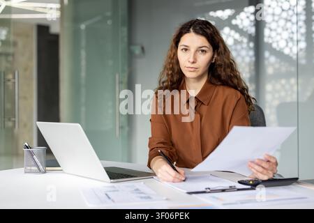 Junge Geschäftsfrau arbeitet an ihrem Schreibtisch, hält Finanzdokumente zur Überprüfung und schreibt Notizen auf einem Klemmbrett mit einem Laptop in der Nähe und konzentriert sich auf tägliche Geschäftsmanagementaufgaben Stockfoto