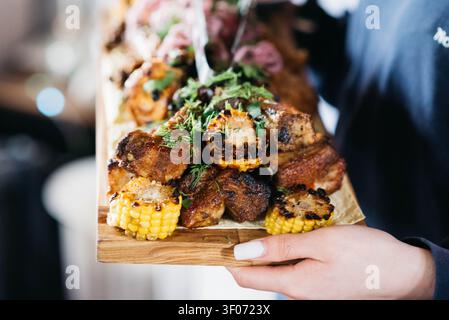 Saftige gegrillte Schweinerippchen, serviert auf einem rustikalen Holzbrett mit verkohltem Mais, frischen Kräutern und Gemüse. Nahaufnahmen von Lebensmitteln mit geringer Schärfentiefe, perfekt für Barbecue-, Restaurant-, Komfortessen- und Gourmetessen-Konzepte. Stockfoto