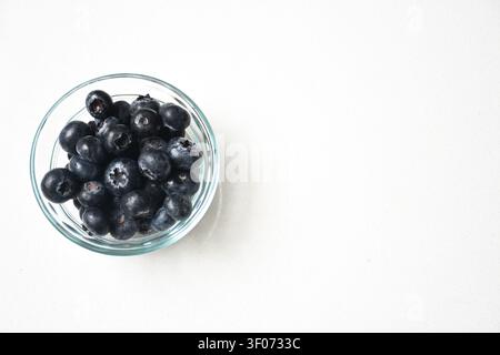 Frische Blaubeeren in einem klaren Glasbehälter, der auf einem sauberen weißen Tisch platziert ist. Minimalistische Komposition mit natürlichem Licht, lebendigen natürlichen Farben Stockfoto