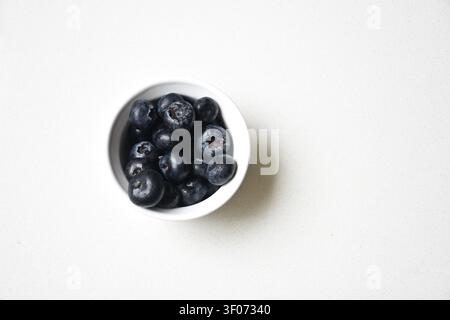 Frische Blaubeeren in einer handgefertigten Keramikschale, die auf einem sauberen weißen Tisch platziert ist. Minimalistische Komposition mit natürlichem Licht, lebendigen Farben Stockfoto