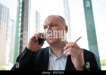 Konzentrierter, reifer Geschäftsmann telefoniert am Smartphone, während er einen Stift in einem modernen Büro hält. Geschäftskommunikation, Verhandlungen und Entscheidungsfindung. Stockfoto