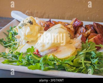 Nahaufnahme eines Gourmet-Brunch-Tellers mit Hummer-Eiern benedict mit Hollandaise-Sauce, gerösteten Kartoffeln und frischem Rucola auf einem weißen Rechteck Stockfoto