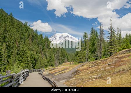 A paved hiking path with a rustic wooden fence winds through a green forest toward a majestic snow-capped mountain peak under a blue sky with white cl Stockfoto