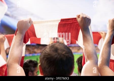 Nahaufnahme eines Fußballfans, der einen rot-weißen Schal über dem Kopf in den Stadionrängen hält. Mann im Teamtrikot hebt den Schal und zeigt während des Live-Spiels seine Unterstützung für das Team Stockfoto