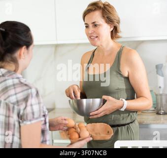Eine Frau mittleren Alters hält eine Schüssel und spricht mit einer anderen Frau in der Küche Stockfoto