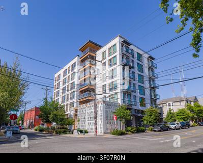 Ein mehrstöckiges weißes Wohngebäude mit Glasbalkonen und dekorativem Metallschirm steht an einer Straßenecke unter klarem blauen Himmel. Stockfoto