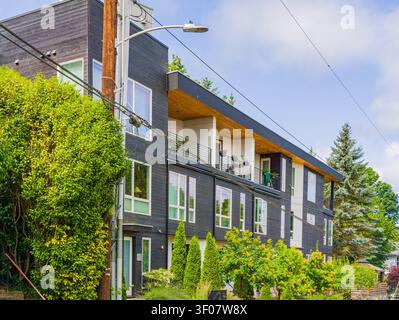 Modernes mehrstöckiges Wohngebäude mit dunkler Abzweigung, weißen Fensterrahmen und einem Holzbalkon, umgeben von üppig grünen Büschen und Bäumen. Stockfoto