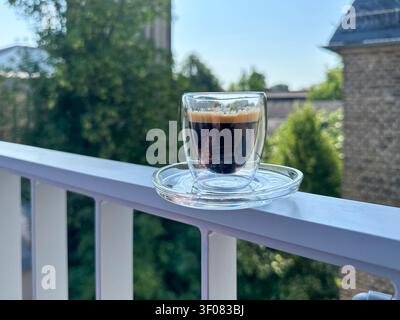 Eine klare Glastasse Espresso steht auf einem weißen Balkongeländer, mit üppiger Vegetation und historischer Ziegelarchitektur im Hintergrund. Stockfoto