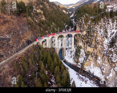 Aus der Vogelperspektive auf das Landwasserviadukt mit dem roten Gletscherexpress, der die Steinbogenbrücke über ein verschneite Tal mit einem Fluss und Kiefern in G überquert Stockfoto