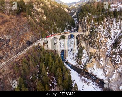 Luftaufnahme des Landwasserviadukts mit einem Zug über die Steinbogenbrücke über eine verschneite Schlucht in Graubünden, Schweiz. Stockfoto