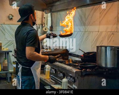 Washington State, USA, 21. Juni 2026 - Ein Küchenchef mit schwarzer Kappe und Handschuhen wirft eine Pfanne mit hohen Flammen in einer Geschäftsküche mit Fliesen Stockfoto