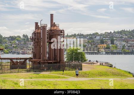 Washington State, JUN 21, 2026 - Verrostete Industrietürme und Schornsteine stehen auf einem grasbewachsenen Hügel mit Blick auf das Wasser und die Stadtsilhouette von Seattle Stockfoto