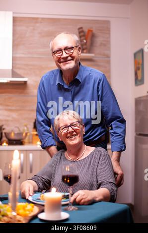 Aufgeregte Frau im Rollstuhl, die mit ihrem Ehemann zu Abend isst, fröhliches, glückliches älteres Ehepaar, das zusammen in der gemütlichen Küche speist, genießt die Mahlzeit, konzentriert auf die aufgeregte Frau im Rollstuhl Stockfoto