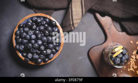 Ansicht von oben mit einer rustikalen Holzschüssel gefüllt mit frischen Blaubeeren neben einem Glas geschichteten Joghurtparfaits, platziert auf einer dunklen strukturierten Oberfläche wi Stockfoto