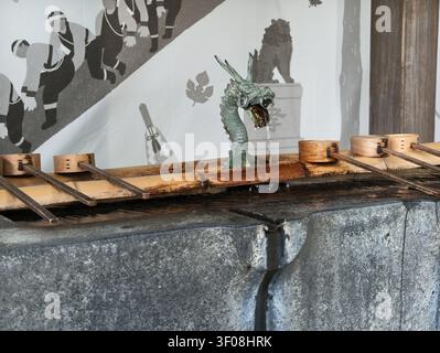 Alte Steinfontäne fließt sanft in das Becken darunter. Metallener Drachenkopf tropft Wasser mit ruhiger Anmut. Holzeimer säumen den Trog, bereit für den Gebrauch. W Stockfoto