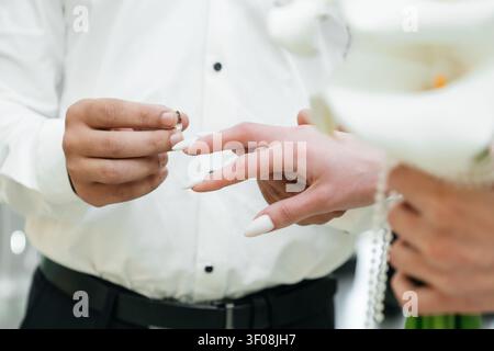 Nahaufnahme eines Bräutigams, der der Braut während der Trauung einen Ehering an den Finger steckt, mit einem Strauß weißer Calla-Lilien Stockfoto