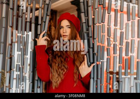 Frauenmode-Porträt eines stilvollen Rothaarigen mit rotem Barett, der einen roten Pullover trägt, hinter einem grauen Perlvorhang im Studio Stockfoto