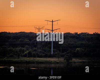 Elektromasten und Übertragungsleitungen, die sich vor einem lebhaften orangefarbenen Sonnenuntergangshimmel über einem Wald und einem reflektierenden Wasserufer abzeichnen Stockfoto
