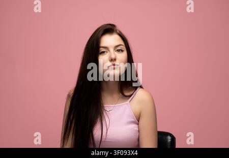 Junge Frau mit ernstem Ausdruck posiert vor einem lebhaften rosa Hintergrund, langes dunkles Haar fällt über die Schultern Stockfoto