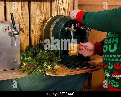Person in festlichem Pullover, die Glühwein aus einem Getränkespender in einen Glasbecher gießt Stockfoto