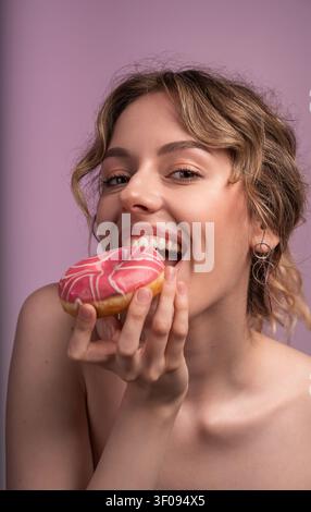 Fröhliche junge Frau beißt in einen appetitlichen rosa glasierten Donut und genießt einen süßen Snack mit einem verspielten Lächeln Stockfoto