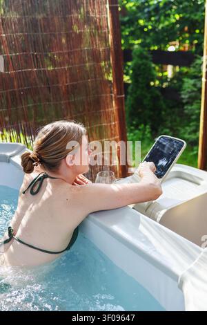 Junge Frau entspannt sich in einem luxuriösen Whirlpool mit Touchscreen-Panel im Garten und schafft ein friedliches Spa- und Selbstpflegeerlebnis. Stockfoto