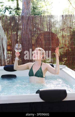 Junge Frau genießt Hydrotherapie in einem Außenspa, spritzt Wasser und hebt ein Glas während eines entspannenden Kurzurlaubs. Stockfoto
