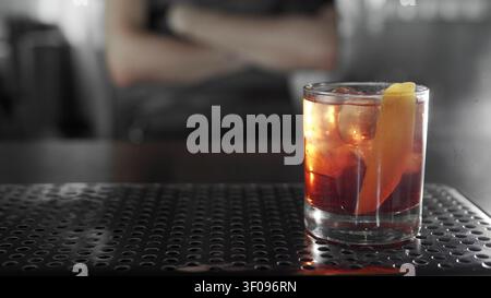 Negroni-Cocktail mit Eis und einer Orangenschale auf einer Metall-Barmatte, Barkeeper im Hintergrund unscharf Stockfoto