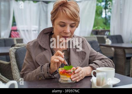 Eine Frau sitzt an einem Tisch in einem Café und isst Dessert aus einem kleinen Glas. Sie schaut konzentriert auf ihr Dessert und hält einen Löffel in der Hand. Eine Tasse Kaffee ist Stockfoto