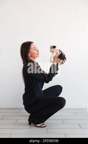 Frau in schwarzem formellen Hosenanzug sitzt an weißer Wand und hält ein kleines süßes Kätzchen. Kaukasisches brünettes Model mit langen Haaren. Geschäftsporträt Stockfoto