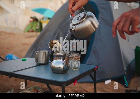 Person gießt heißes Wasser aus einem Edelstahl-Wasserkocher in eine Teekanne auf einem Campingtisch neben einem Zelt am Strand, was entspanntes Camping im Freien und morgendliche Rituale vermittelt Stockfoto