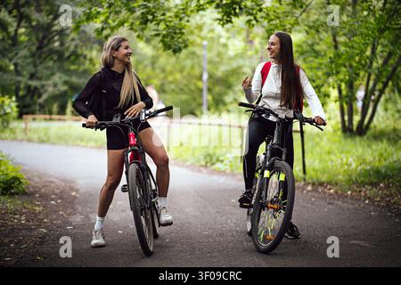 Zwei glückliche Frauen mit Mountainbikes lachen und unterhalten sich auf einem gepflasterten Waldweg während eines sonnigen Outdoor-Fahrradabenteuers. Stockfoto