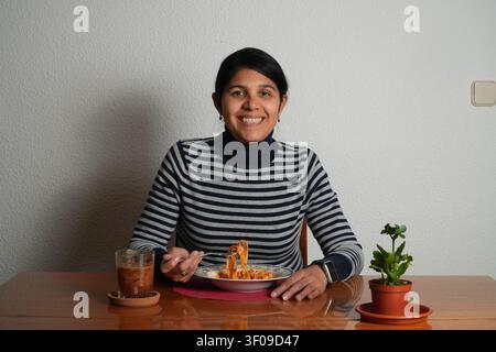 Frau lächelt in die Kamera, hält einen Teller mit Essen. Ein Glas mit dunklem Getränk steht neben ihr. Eine kleine Topfpflanze steht auf dem Tisch in ihrer Nähe rechts. Einfach, rusti Stockfoto